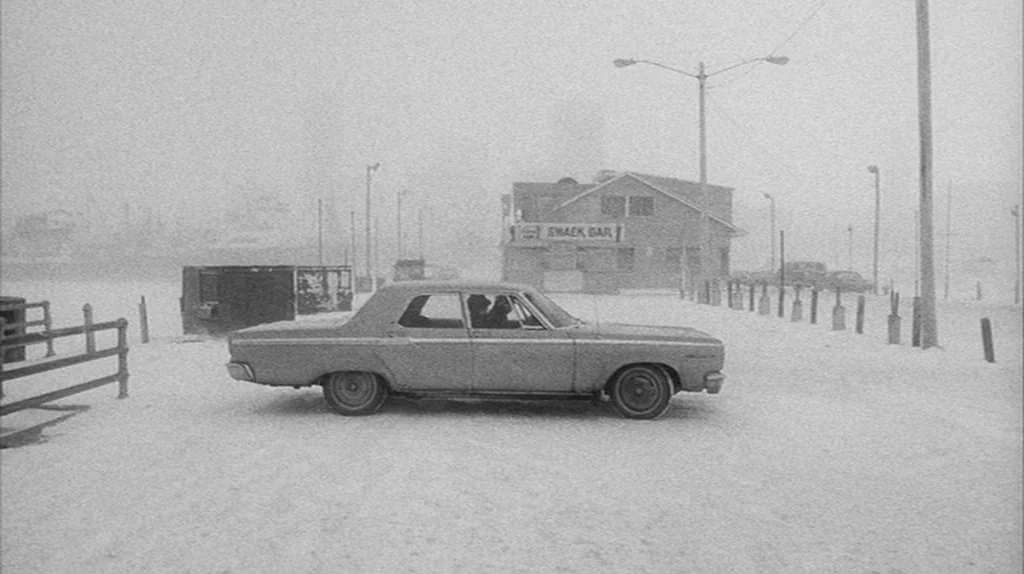 Stranger Than Paradise (1984) movie still: wide — A snow-covered parking lot surrounds a lone sedan in front of a snack bar. Falling snow obscures…