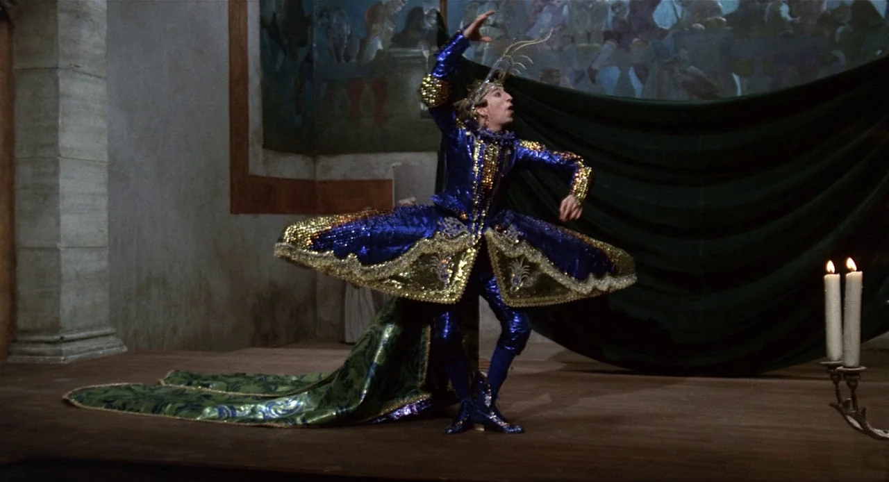 Fellini’s Casanova (1976) movie still: fullbody — A stone-walled hall features green drapery and a painted mural in the background. A male dancer in…