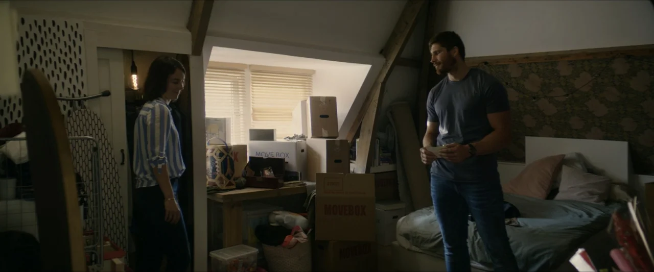 Black Lotus (2023) movie still: fullbody — An attic bedroom with slanted ceiling beams contains several cardboard packing boxes and a bed. A man and…