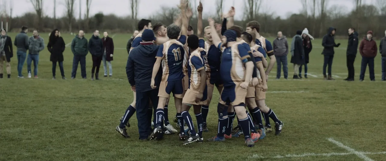 The Thorn On His Side (2014) movie still: fullbody — A group of rugby players huddled together on a grassy field in overcast weather. The players in…