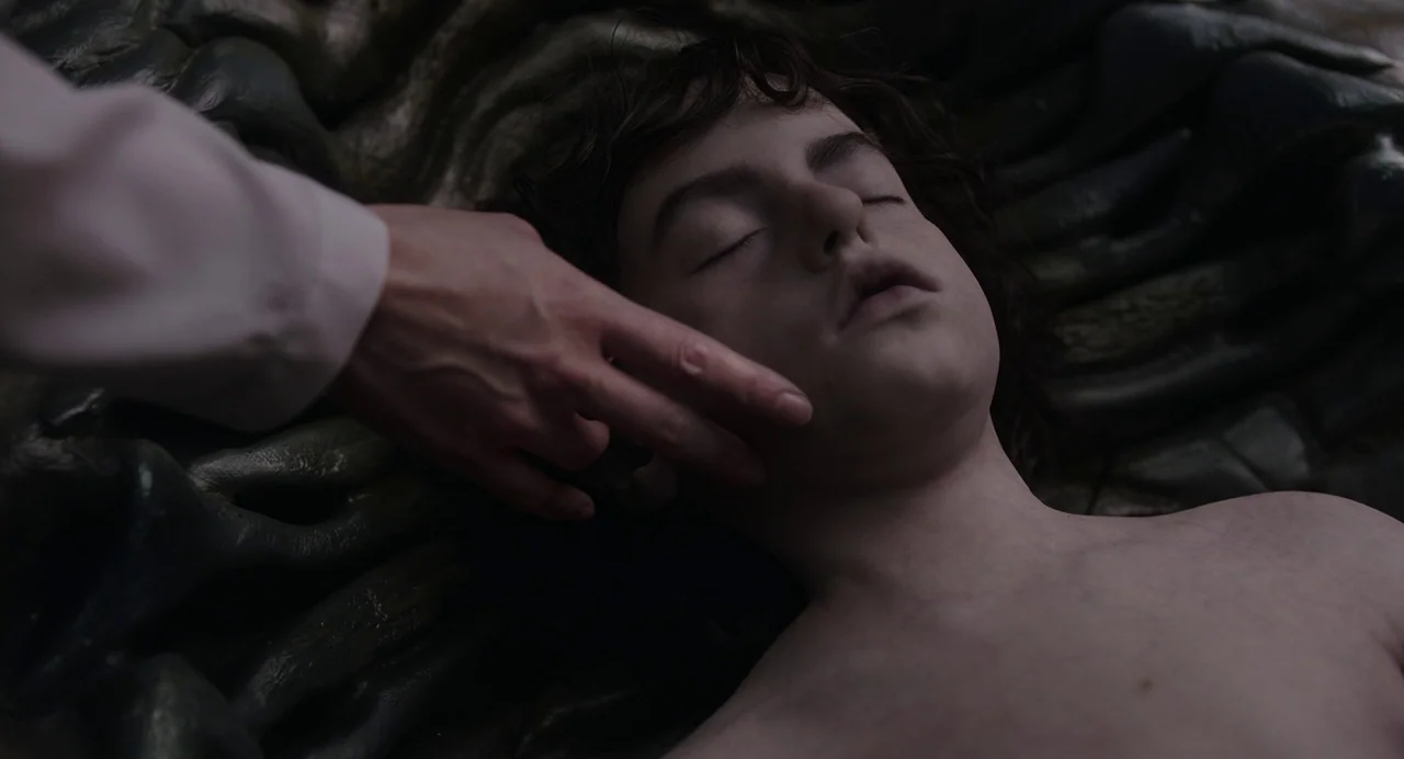 Crimes of the Future (2022) movie still: closeup — A dark, textured organic environment surrounds a young man lying down with eyes closed. Another…