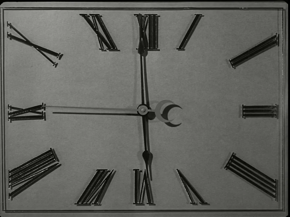 Modern Times (1936) movie still: closeup — A rectangular analog clock face displays stylized Roman numerals against a plain gray background. Three…