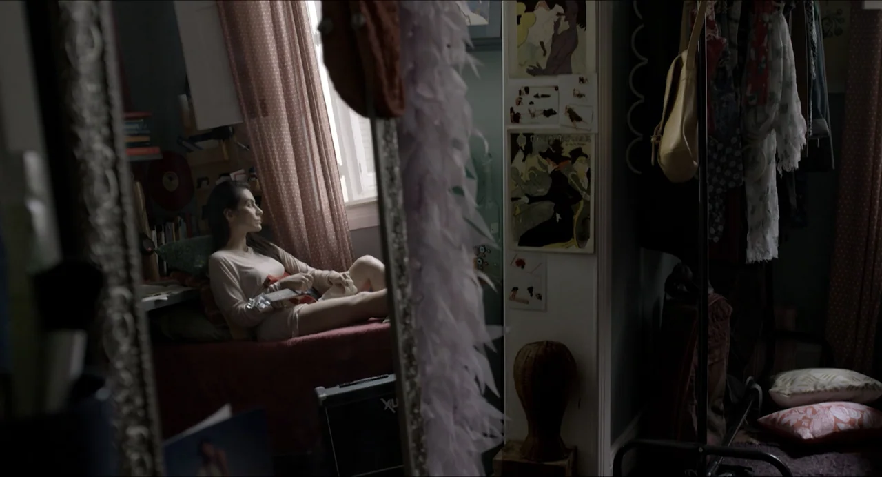 Suspension Points (2022) movie still: medium — A cluttered bedroom is filled with clothing racks, posters, and personal items. A woman sits on a bed…