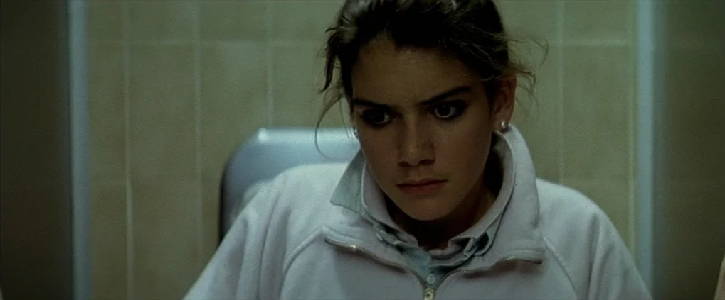 Anguish (1987) movie still: closeup — A tiled restroom interior features walls covered in pale green rectangular tiles. A young woman stands…