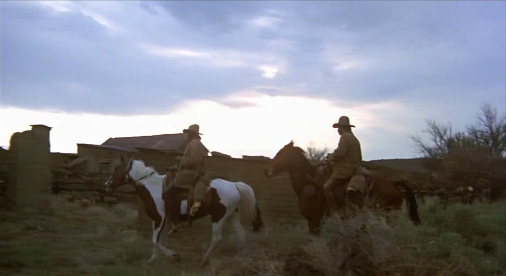 The Hired Hand (1971) movie still: wide — Two men on horseback ride past a dilapidated, low-profile wooden structure at twilight. The sky is…