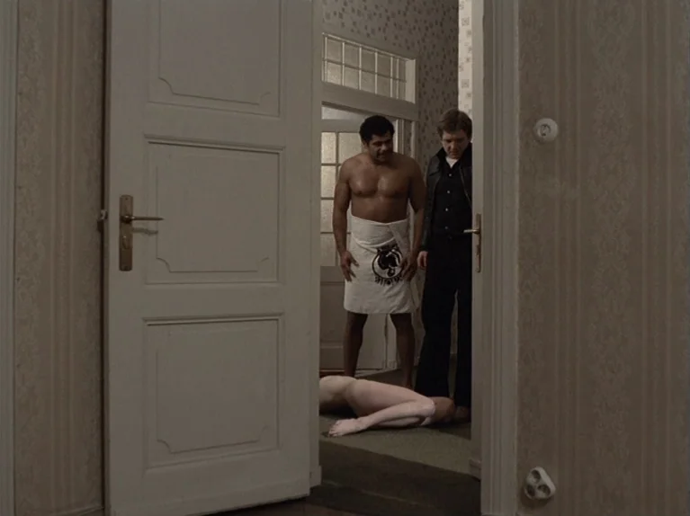 The Third Generation (1979) movie still: medium — A residential hallway with patterned beige wallpaper and a white wooden door frame. Two men stand…