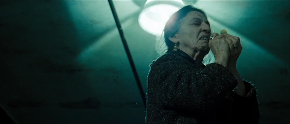 Drag Me to Hell (2009) movie still: medium — A dimly lit, concrete basement space features a single industrial light fixture above. An elderly woman…