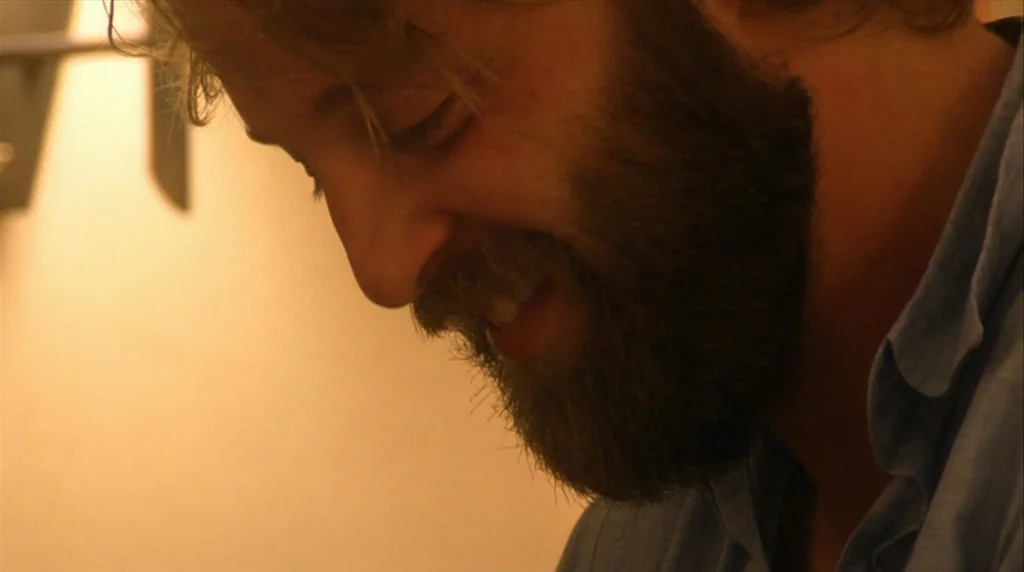 Humpday (2009) movie still: closeup — A man with a full beard looks downward inside a dimly lit interior space. His face is illuminated from the…
