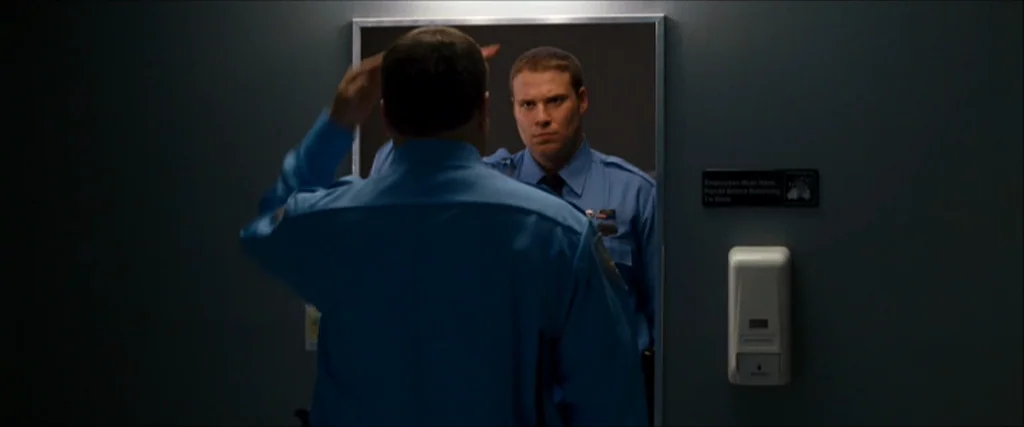 Observe And Report (2009) movie still: medium — A public restroom interior with grey walls and a mirror mounted above a dispenser. A man in a blue…