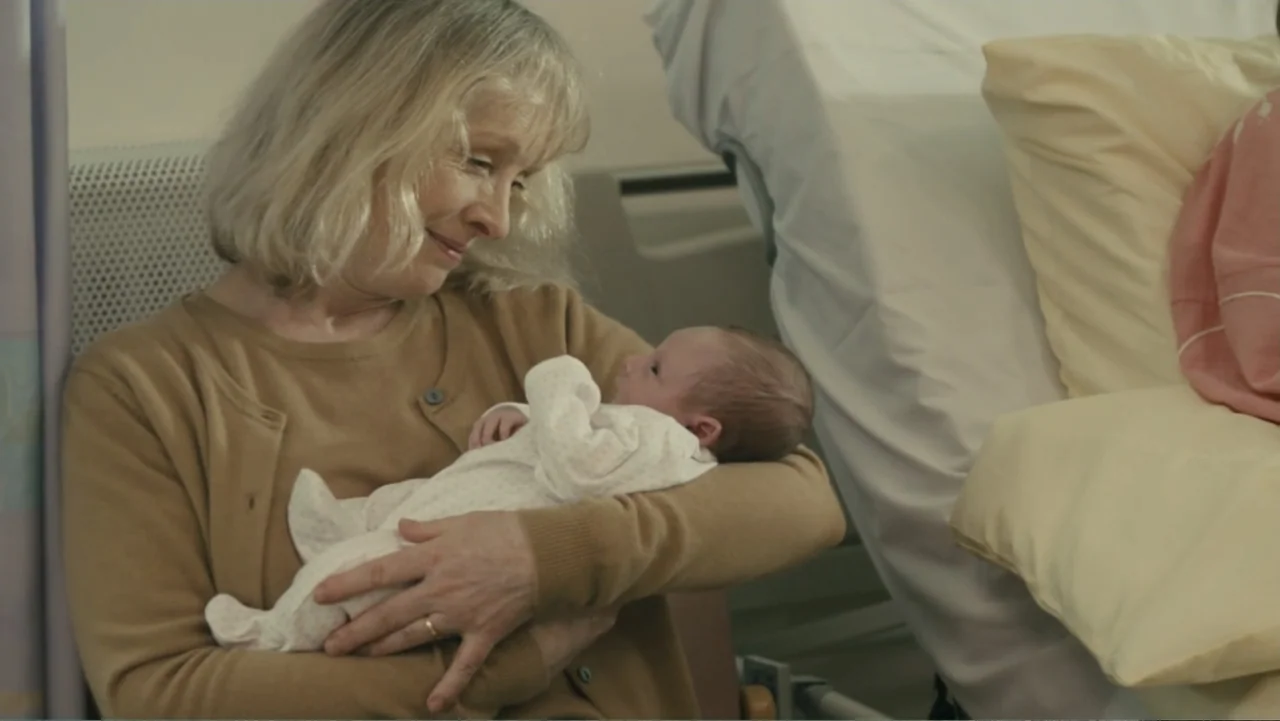 About Time (2013) movie still: medium — A hospital room with a light-colored wall and a hospital bed behind the subject. A woman in a tan cardigan…