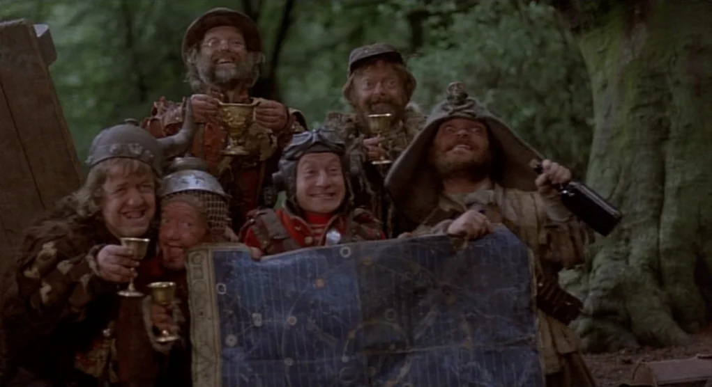 Time Bandits (1981) movie still: medium — A group of six small men stand together in a wooded area with trees. The men hold up a large, blue…