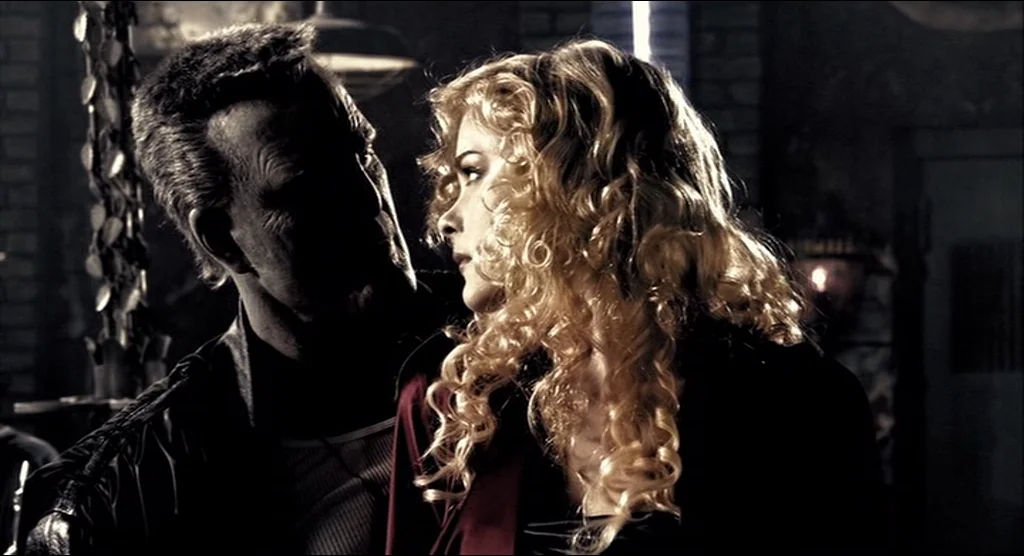 Sin City (2005) movie still: medium — A dimly lit room features a high-contrast environment with deep shadows and texture. A man and a woman stand…