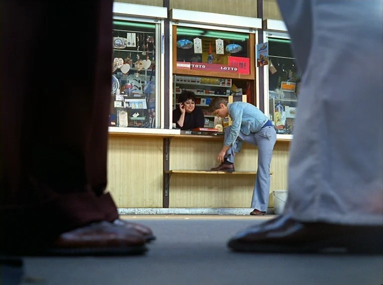 Fox and His Friends (1975) movie still: wide — A street-side lottery kiosk with glass windows filled with small trinkets and signs. A young man ties…