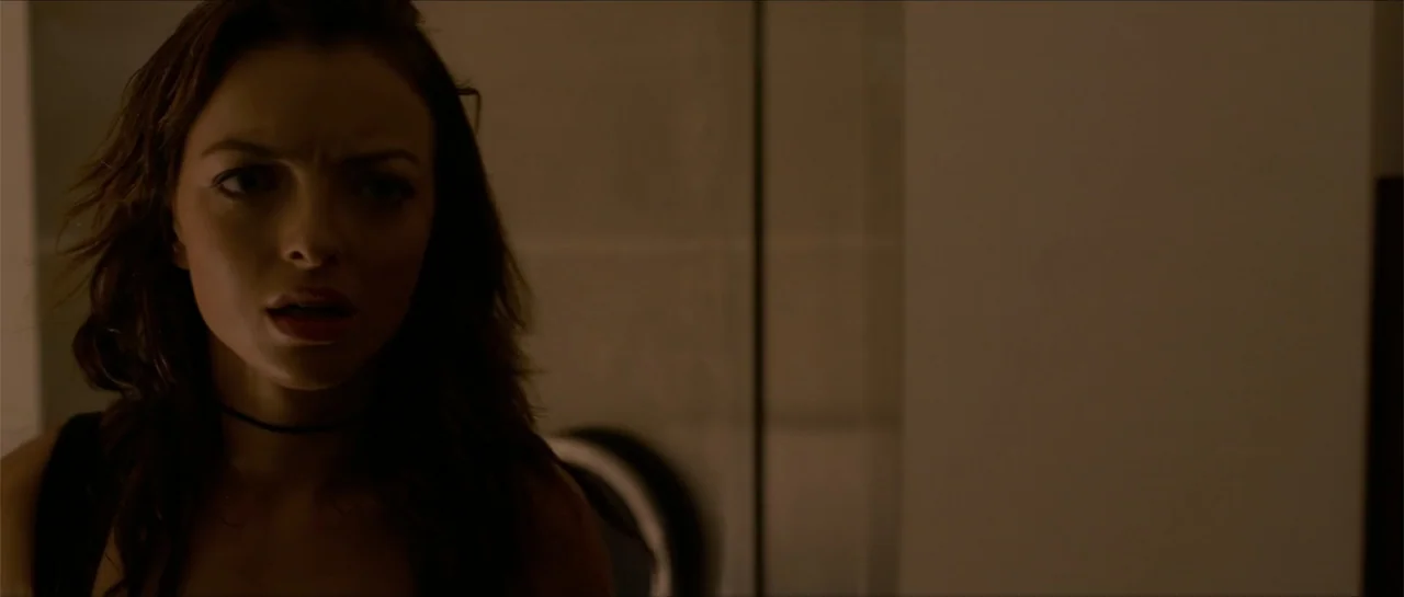 The Vault (2017) movie still: closeup — A dimly lit interior space with beige tiled walls. A young woman with dark hair turns her head to look…