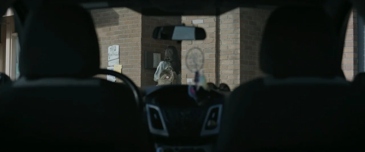 It Feeds (2025) movie still: medium — The view from inside a car looks through the windshield toward a brick building entrance. A woman with a…