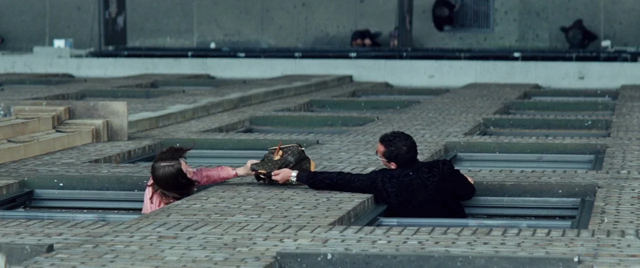 Uncut Gems (2019) movie still: wide — A flat rooftop covered with repeating brick skylights features two people at openings. They reach across a…