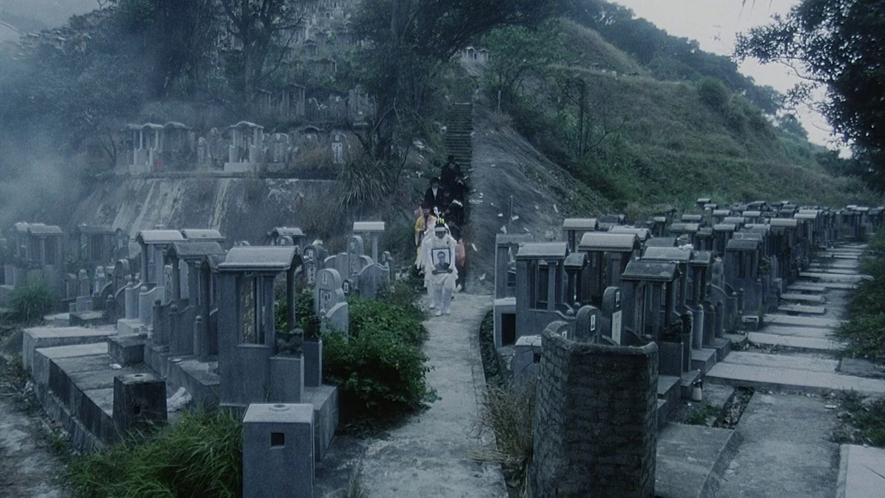 Visible Secret (2001) movie still: wide — A hillside cemetery features rows of stone markers lining a narrow paved pathway. A group of people…