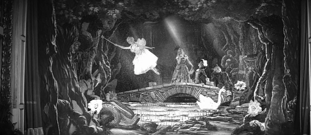 The Elephant Man (1980) movie still: wide — A theatrical stage set features a painted forest backdrop, a stone bridge, and a pond. A woman dressed…