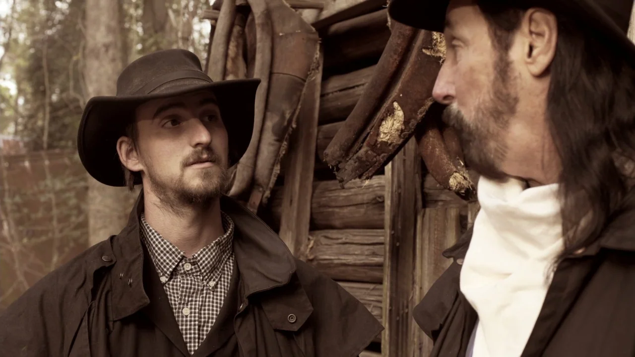 The Dalton Gang (2020) movie still: medium — Two men in 19th century attire stand in front of a rustic log wall. The younger man faces the older man…