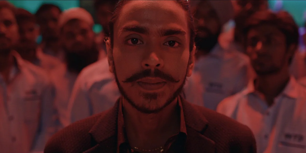 The White Tiger (2021) movie still: closeup — A man stands prominently in front of a blurred crowd inside a dim facility. He stares directly ahead…