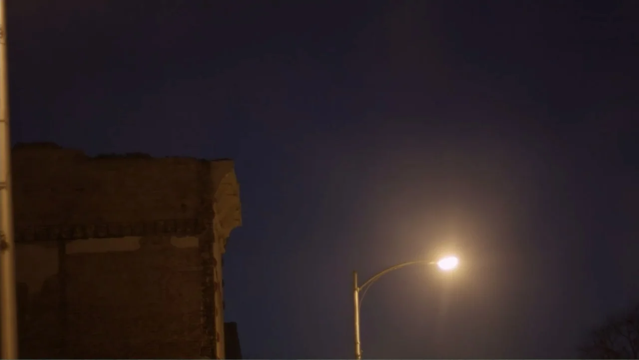 Rich Hill (2014) movie still: establishing — A dark urban scene features the top corner of a weathered brick building silhouette. A single…