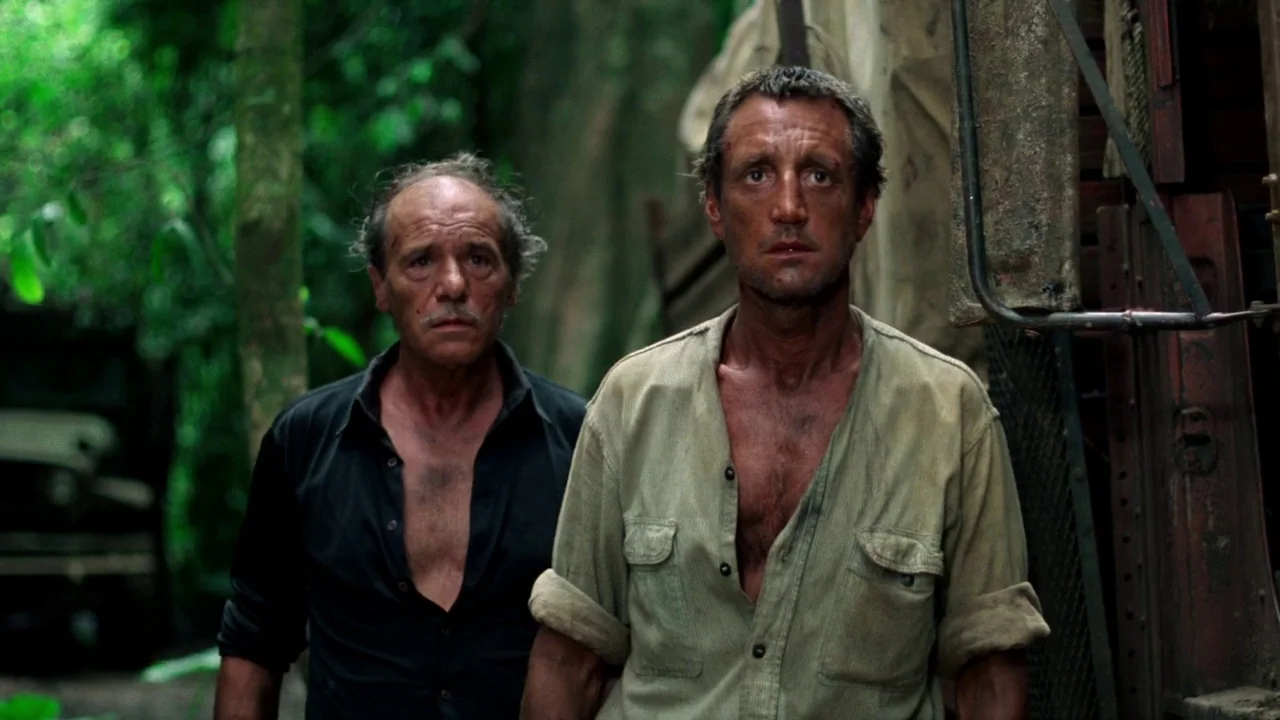 Sorcerer (1977) movie still: medium — Two men stand closely together in a dense, green, overgrown tropical jungle environment. Both men appear…