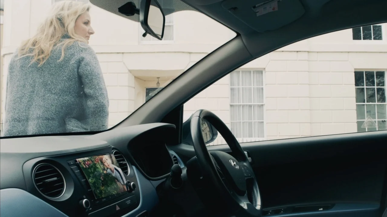 You Might Get Lost (2021) movie still: medium — A view from inside a car looking out at a suburban residential building exterior. A blonde woman in…