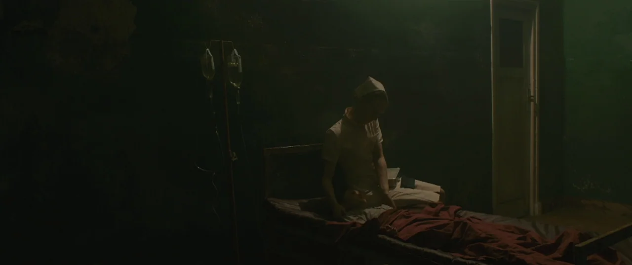 Evolution (2015) movie still: medium — A dark hospital ward has a single bed with a patient and a nurse. The nurse sits on the edge of the bed with…