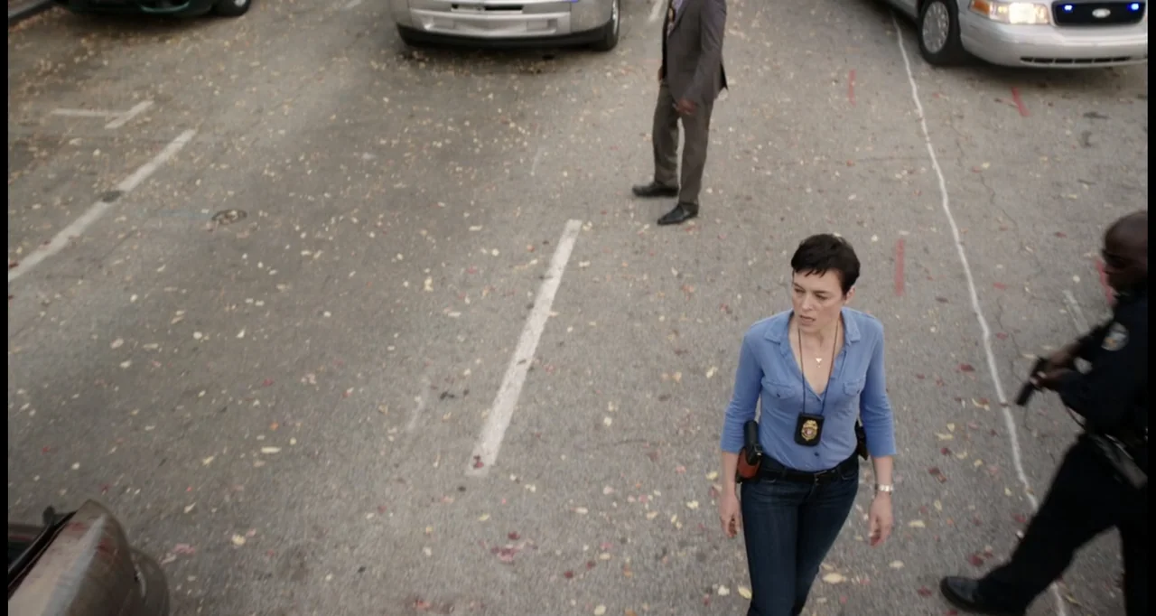 Sabotage (2014) movie still: medium — An asphalt parking lot scattered with fallen leaves contains several parked police vehicles. A female…