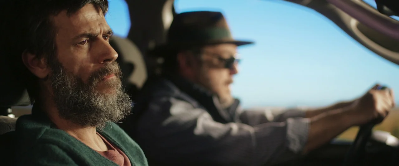 When Evil Lurks (2023) movie still: medium — Interior of a moving car with two men sitting in the front seats. The passenger looks ahead while the…