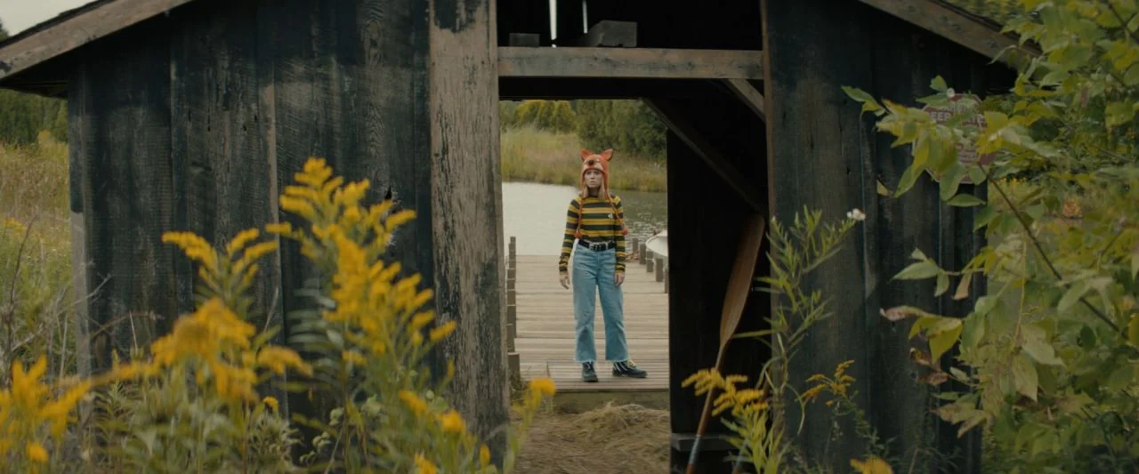 Becky (2020) movie still: fullbody — A weathered dark wood shed sits at the edge of a calm lake. A young woman wearing an animal-eared hat stands…
