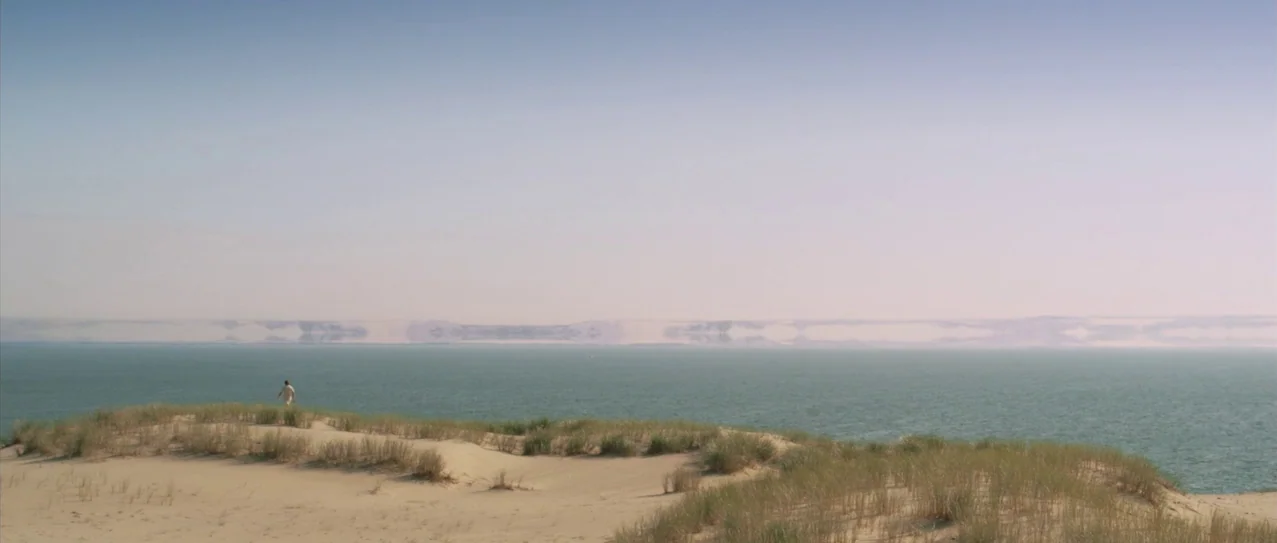 Vanishing Waves (2012) movie still: wide — A vast sandy dune covered with sparse tall grass overlooks a large body of water. A single small figure…