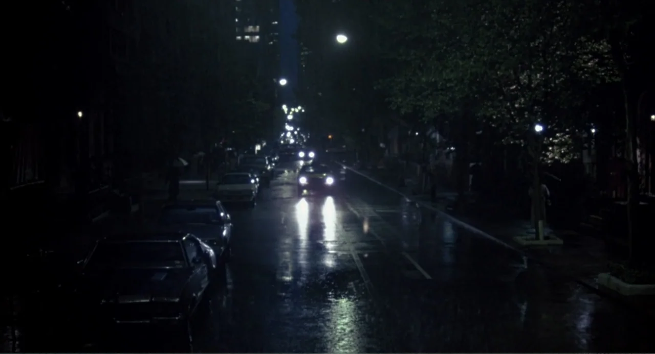 F/X (1986) movie still: establishing — A dark city street at night is slick with rain and reflections. Several parked vintage cars line the curbs as…