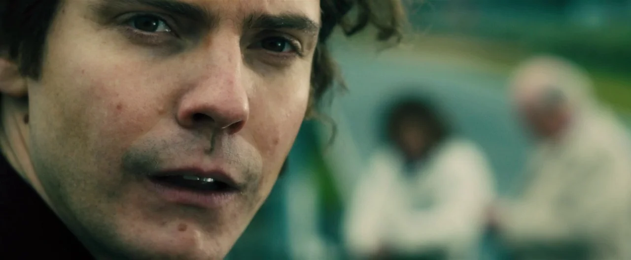 Rush (2013) movie still: closeup — An outdoor area with blurred figures visible in the out of focus background. A man looks directly toward the…