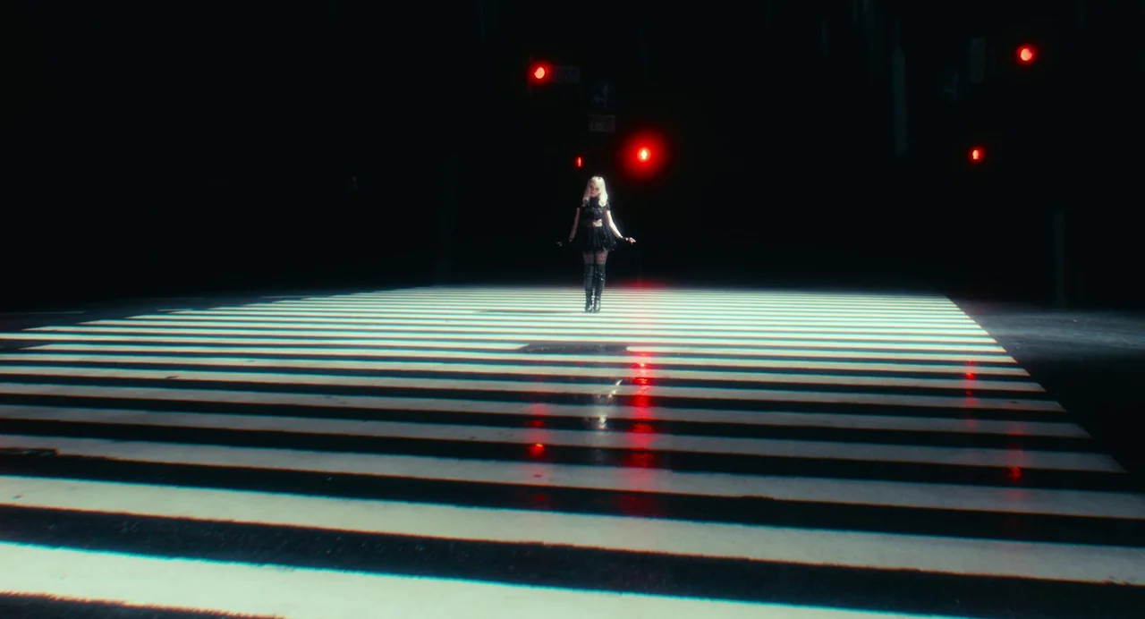 xxxHolic (2022) movie still: wide — A dark urban intersection at night features a brightly lit zebra crossing. A lone woman stands centered in the…