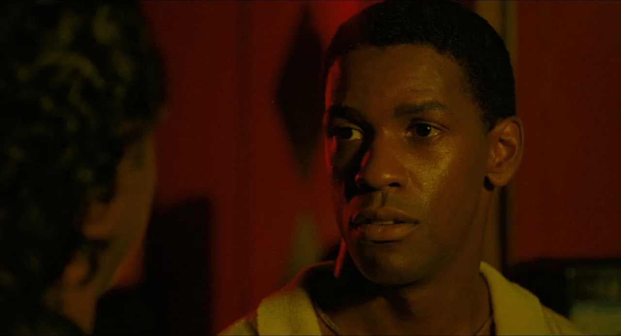 Mississippi Masala (1991) movie still: closeup — A dimly lit interior space features dark red walls. A young Black man looks toward an unseen…