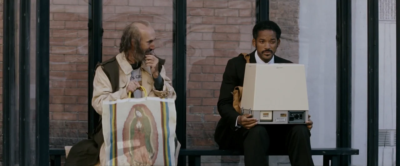 The Pursuit of Happyness (2006) movie still: medium — A glass and brick bus shelter stands on a city sidewalk during the day. Two men sit on a…