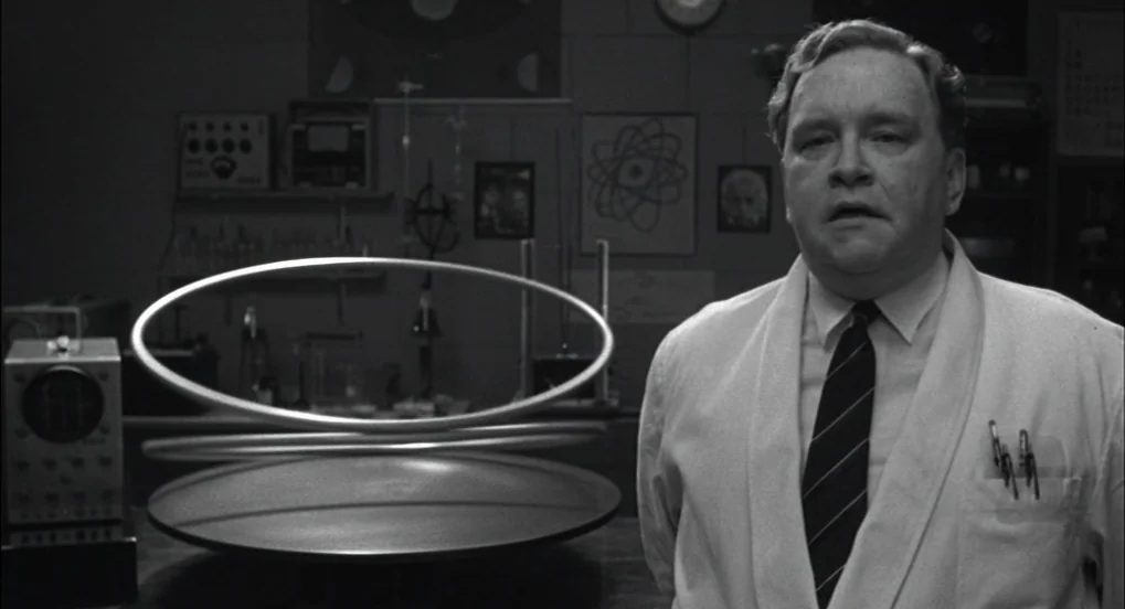 The Hudsucker Proxy (1994) movie still: medium — A 1950s laboratory features scientific diagrams and electronic instruments against a tiled wall. A…