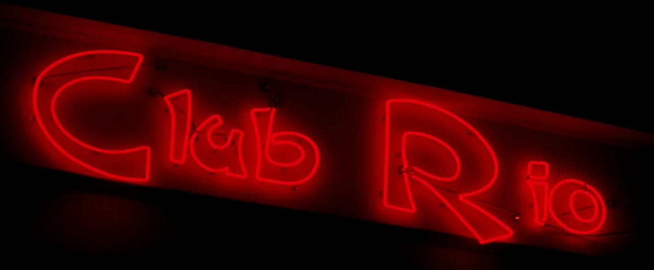 The Yards (2000) movie still: closeup — A tilted neon sign displaying the words Club Rio in bright red letters. The sign is set against a dark,…