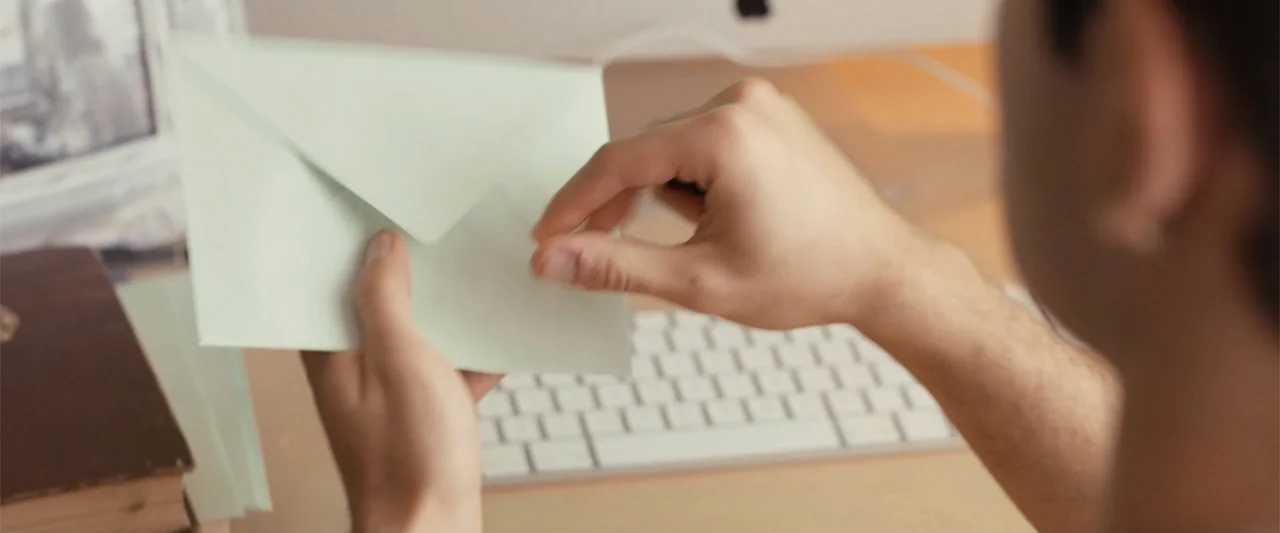 Wrong Package (2025) movie still: closeup — A workspace with a white computer keyboard and a stack of books. A person opens a light green paper…