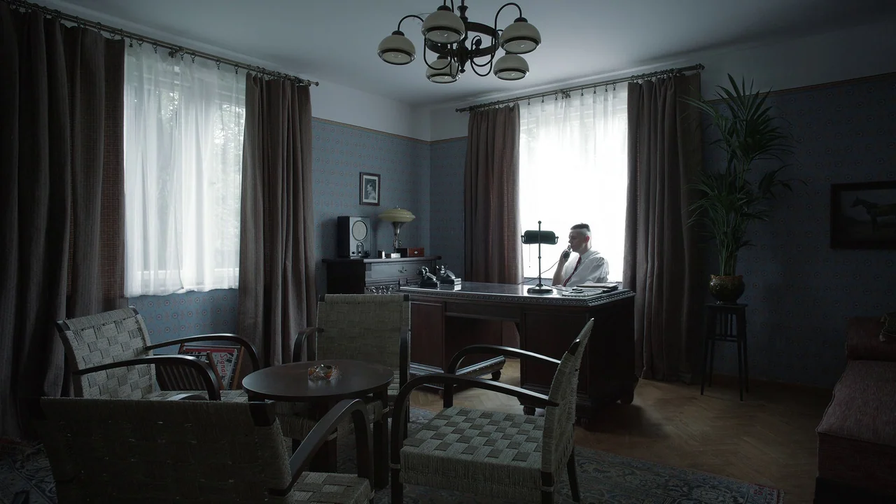 The Zone Of Interest (2023) movie still: wide — A 1940s style room features patterned blue wallpaper and dark wood furniture throughout. A man sits…