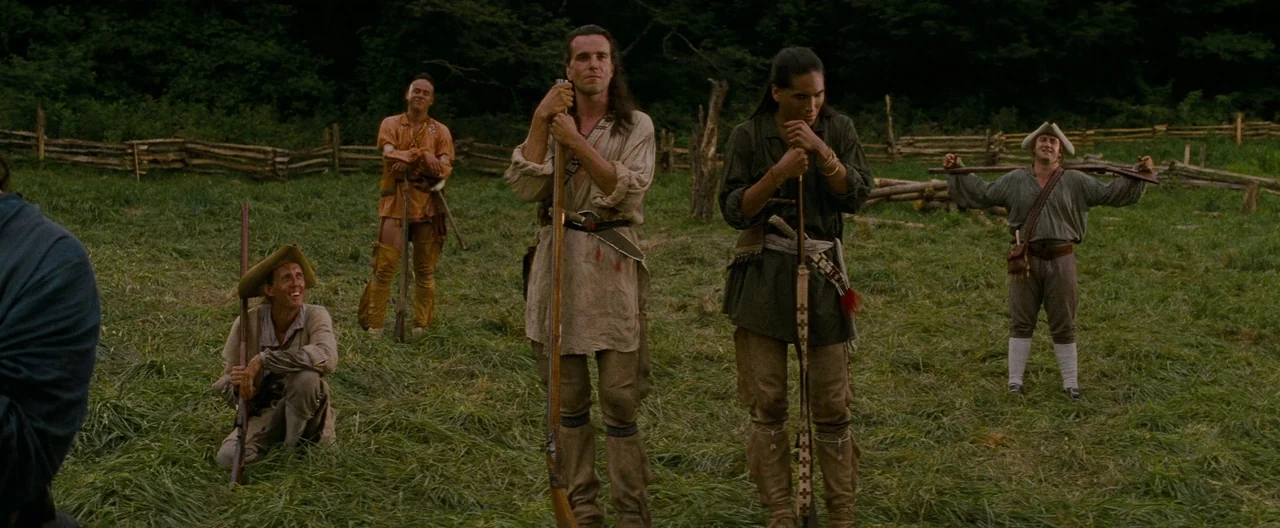 The Last of the Mohicans (1992) movie still: fullbody — A grassy meadow surrounded by a wooden rail fence and dense dark forest. Five men stand…