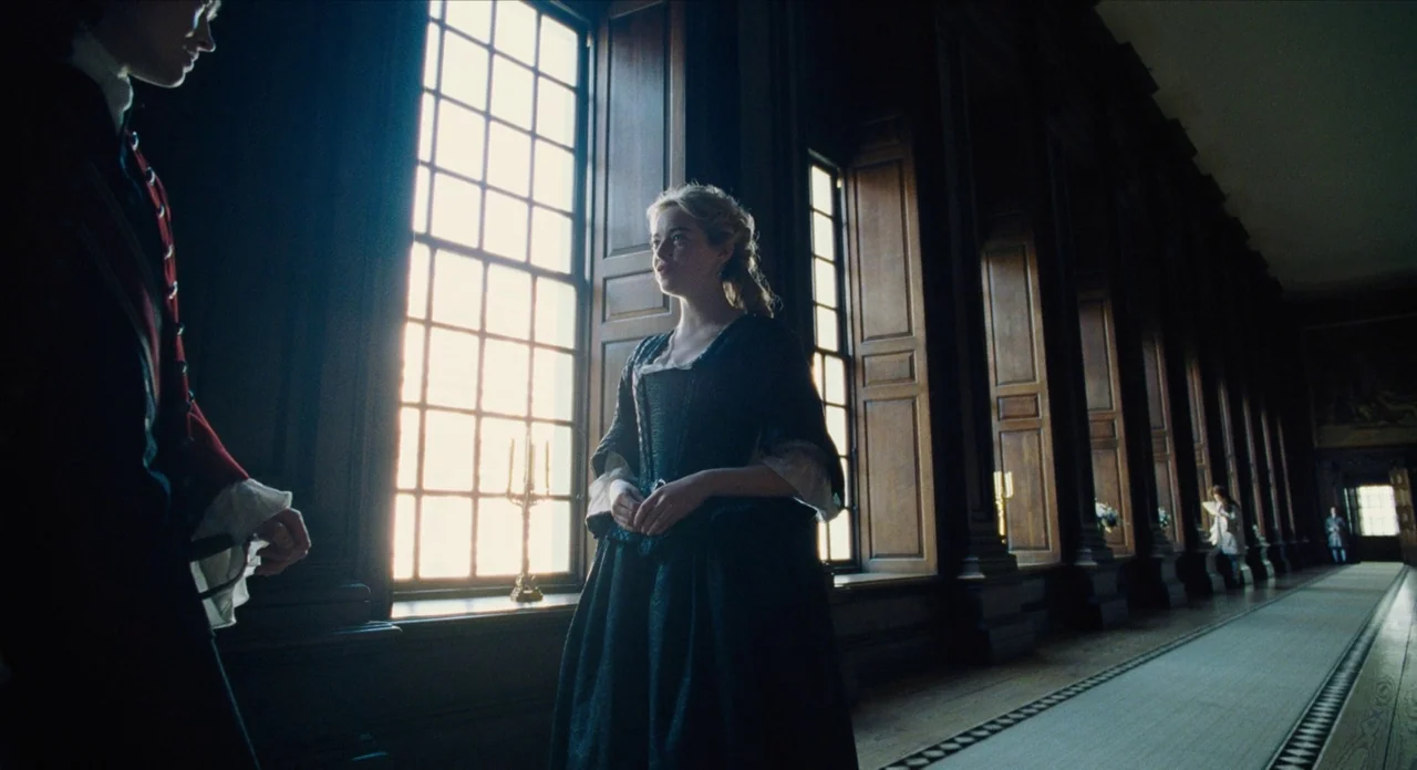 The Favourite (2018) movie still: fullbody — A long, dark wood-paneled gallery features a row of tall windows and a runner. A woman stands in front…
