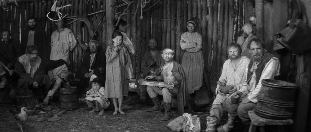 Andrei Rublev (1966) movie still: wide — A rustic, log-walled structure contains several villagers sitting on hay or wooden stools. The group…