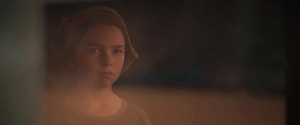 Settlers (2021) movie still: closeup — A dark, dimly lit interior space with the subject centered against a blurred background. A young child…