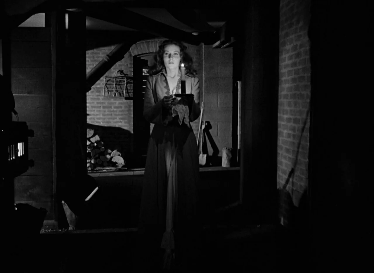 The Spiral Staircase (1946) movie still: fullbody — A dimly lit stone cellar with brick walls and wooden beams overhead. A woman holding a single…