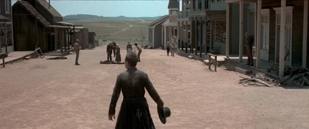 In A Valley of Violence (2016) movie still: wide — A dusty unpaved road runs between wooden buildings in a sparsely populated arid town. A man in a…
