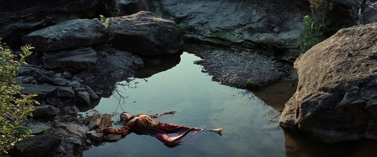 Killers of the Flower Moon (2023) movie still: wide — A quiet rocky creek with dark still water surrounded by large grey boulders. A man lies face…