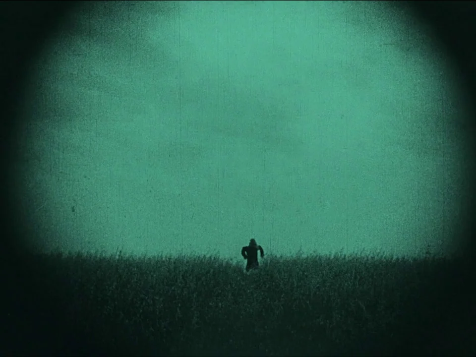 Nosferatu (1922) movie still: wide — A vast, open field extends toward a cloudy, monochromatic teal sky at night. A lone figure in a long coat runs…