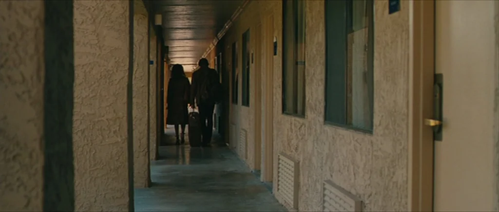 Away We Go (2009) movie still: wide — A long, concrete outdoor motel walkway with textured beige stucco walls and dark windows. A man and a woman…